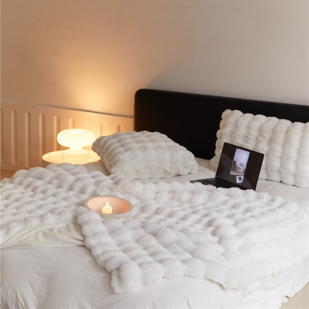 A close-up shot of a white faux rabbit fur throw blanket with a bubble pattern, placed on a bed with a matching pillow.