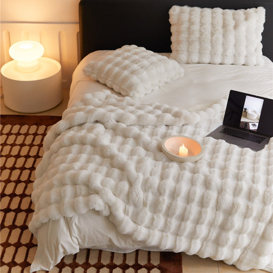 A cozy, white faux rabbit fur throw blanket with a bubbly texture, draped over a bed. A matching pillow and a laptop are on the bed.