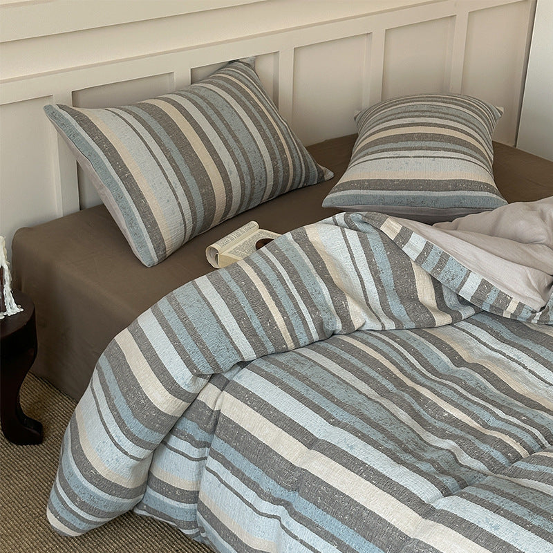 Striped cotton gauze bedding set in soft blue, gray, and beige tones on a brown fitted sheet with white paneled headboard