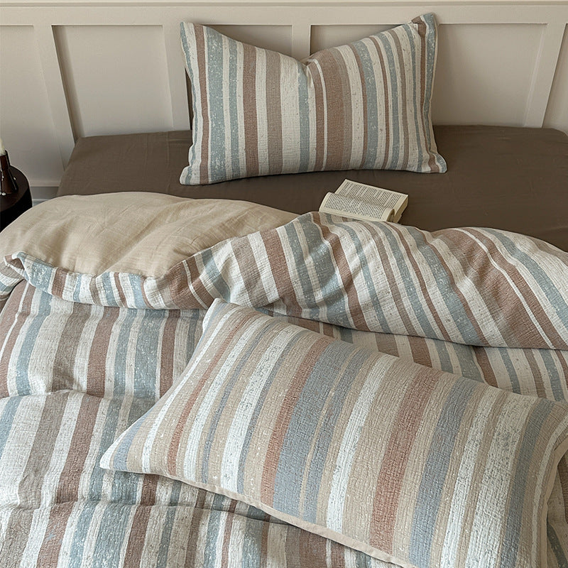 Striped cotton gauze bedding set with blue, beige, and white tones on a brown fitted sheet, open book on bed