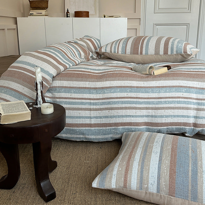 Cozy bed with striped cotton gauze bedding set in earthy tones, featuring pillows and a small wooden side table with candles and a book