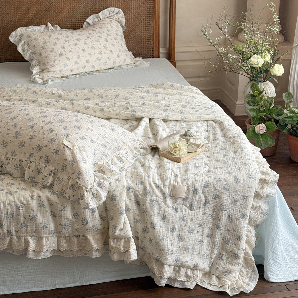 Full view of a bed featuring the Blue Rose Comforter, a ruffled cotton gauze quilt, laid over a light blue bed sheet, emphasizing the charming cottage style, rattan headboard, and floral bedding.