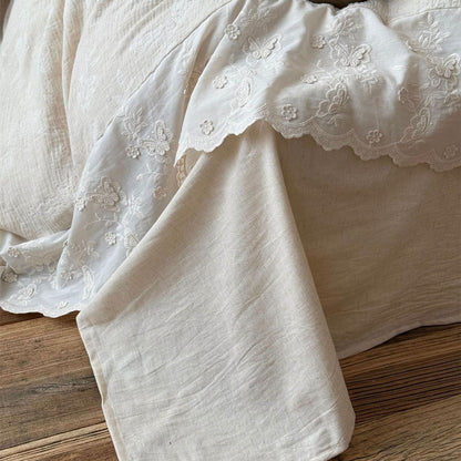 Close-up of textured beige linen cotton bedding with white embroidered floral and butterfly trim on wooden floor