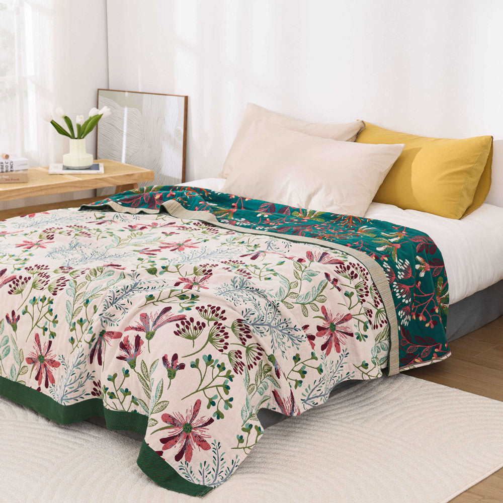 A cream-colored vintage floral throw blanket with burgundy and green botanical prints draped over a bed with white linens.