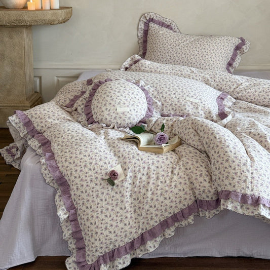 Vintage cottage style purple floral bedding set styled on a low bed, featuring a ditsy floral pattern quilt with intricate purple ruffled lace trim and a matching round cushion.
