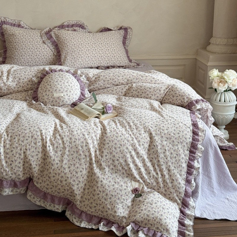 A cozy bedroom setup showcasing the purple floral lace duvet cover set from a side view, styled with a stone pillar vase filled with white roses next to the bed.