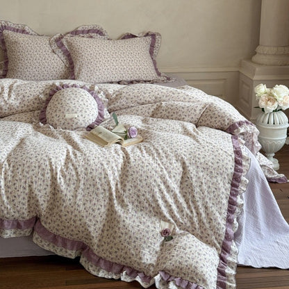 A cozy bedroom setup showcasing the purple floral lace duvet cover set from a side view, styled with a stone pillar vase filled with white roses next to the bed.