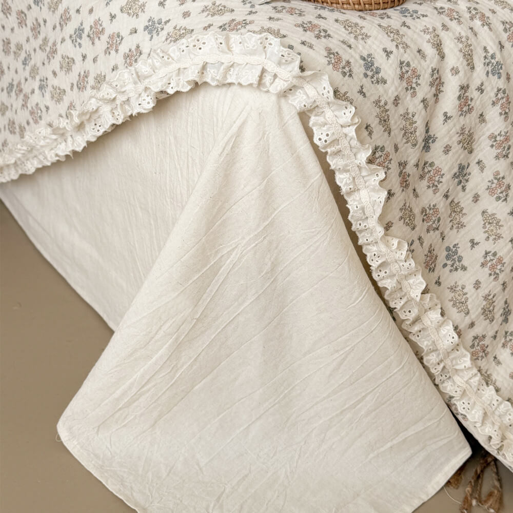 Detailed close-up of a cream-colored linen bed skirt underneath a draped vintage floral summer quilt with lace borders.