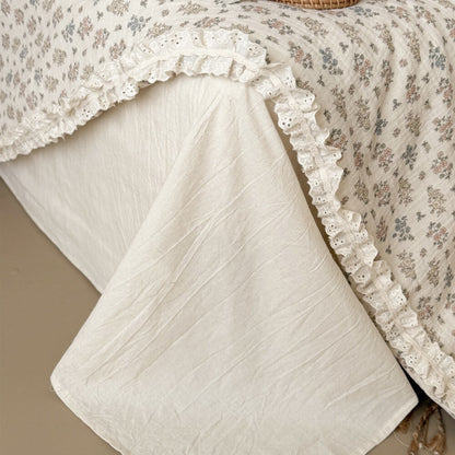 Detailed close-up of a cream-colored linen bed skirt underneath a draped vintage floral summer quilt with lace borders.