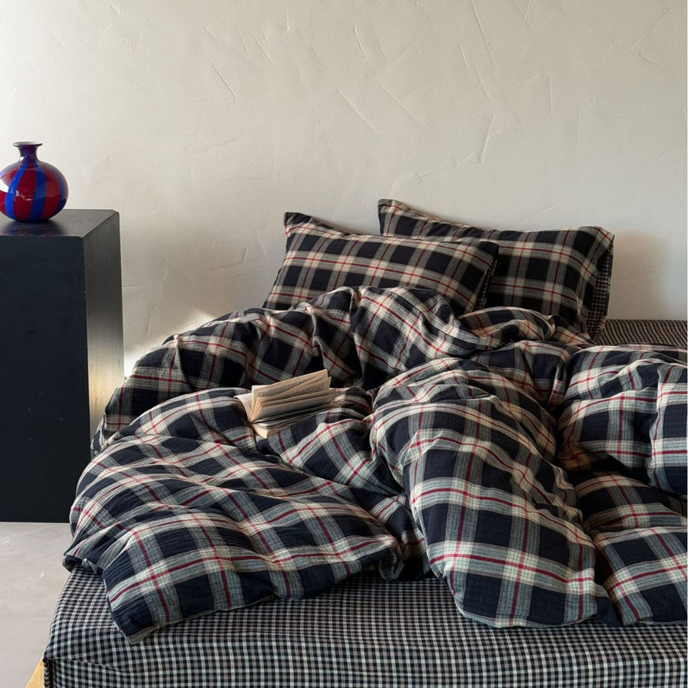 Front view of a ruffled dark plaid cotton muslin bedding set with a matching small-checkered sheet, styled with a glass vase in a minimalist room.