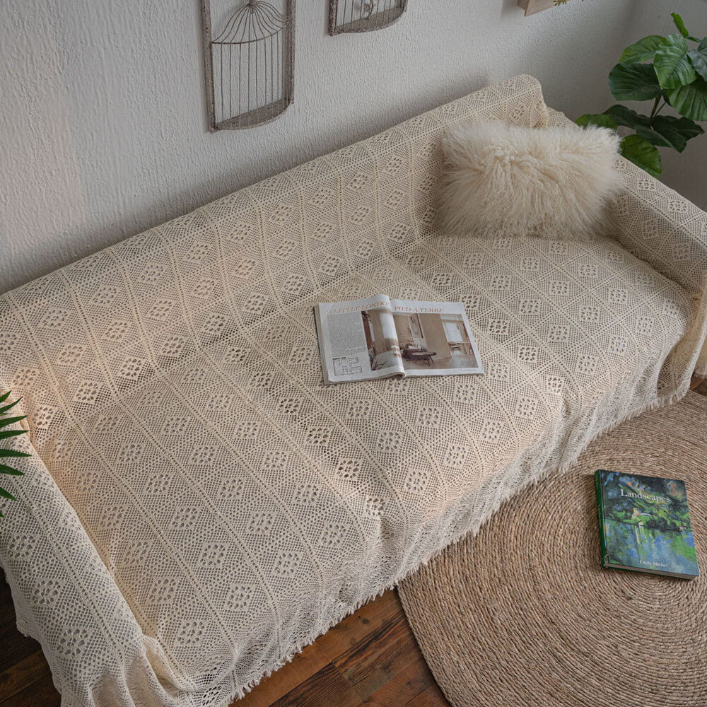 Vintage crochet lace sofa cover with tassel trim on a couch, cozy pillow, and magazines in a natural home setting.