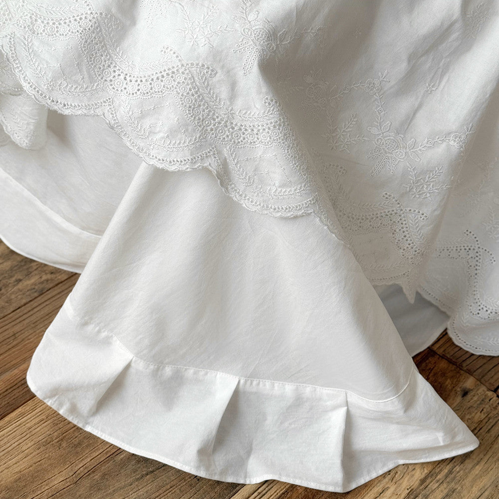 White cotton bedding set with ruffled trim and delicate floral embroidery on wood floor