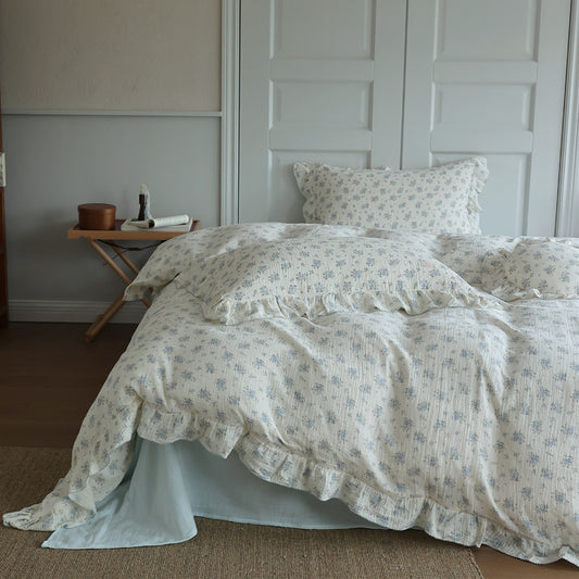 A side view of a bed with a blue floral duvet cover set. The cotton muslin bedding is casually draped, highlighting the long ruffled edges. 