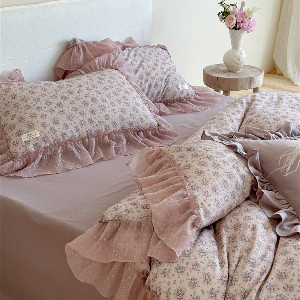 Lavender floral ruffled bedding in a bedroom scene: The bed is dressed with layers of floral pillows, with a small wooden side table holding flowers nearby, creating a romantic and cozy bedroom atmosphere.