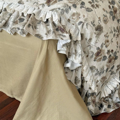 Close-up of lace-trimmed floral bedding showing muted olive flowers, scalloped cutwork lace, ruffled edge, and candle styling