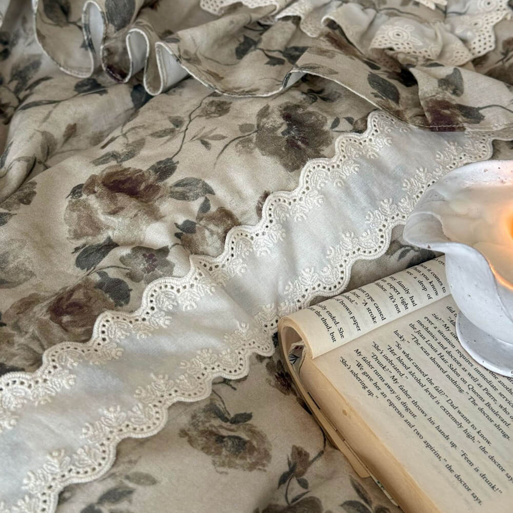 Close-up of vintage floral cotton muslin bedding showing ivory cutwork lace trim, muted hand-painted flowers, and ruffled fabric detail