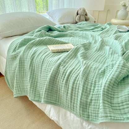 A soft sage green 6-layer cotton gauze blanket laid out as a throw on a bed in a sunny room.